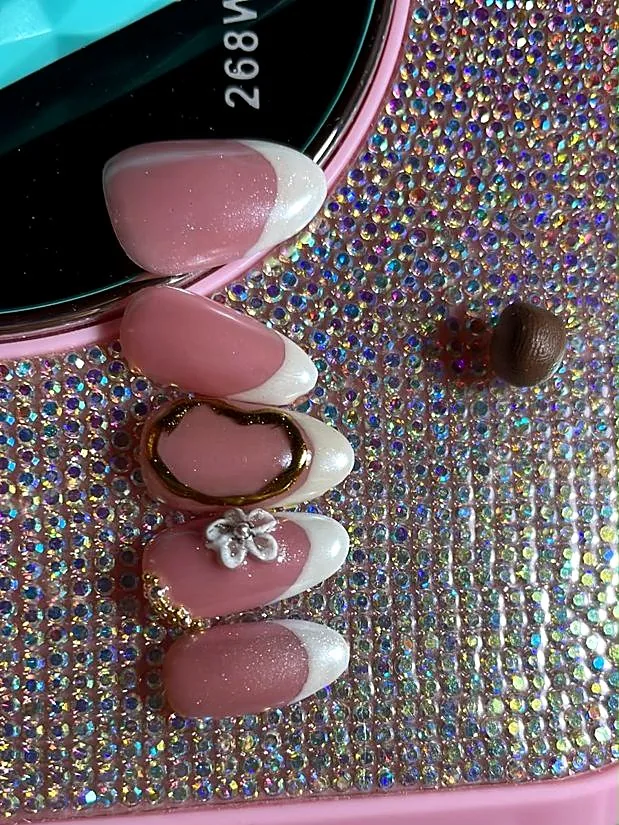 Pink almond French nails with white tips, a gold oval and a 3D flower, resting on a crystal base