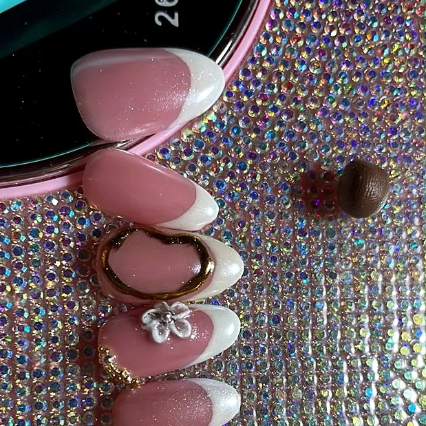 Pink almond French nails with white tips, a gold oval and a 3D flower, resting on a crystal base