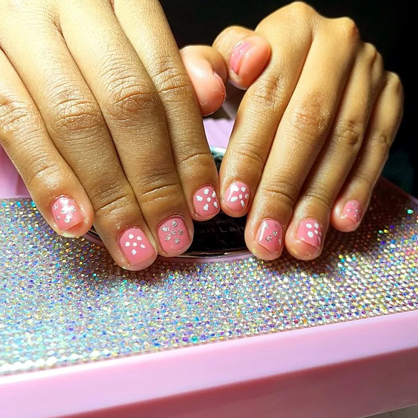 Hands with short pink nails decorated with white dots and glitter
